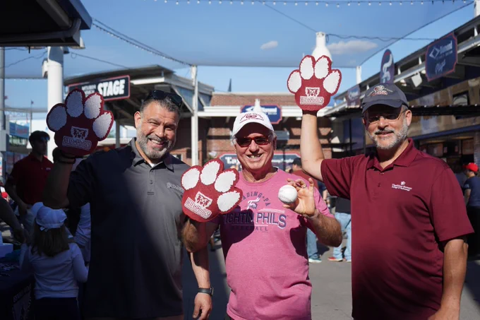 Three men at an outdoor event wearing large red foam paw gloves, one holding a baseball, with a crowd and booths in the background.