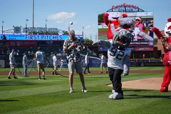 Mascot wearing number 92 jersey throws a baseball on baseball field near photographers and scoreboard at FirstEnergy Stadium.
