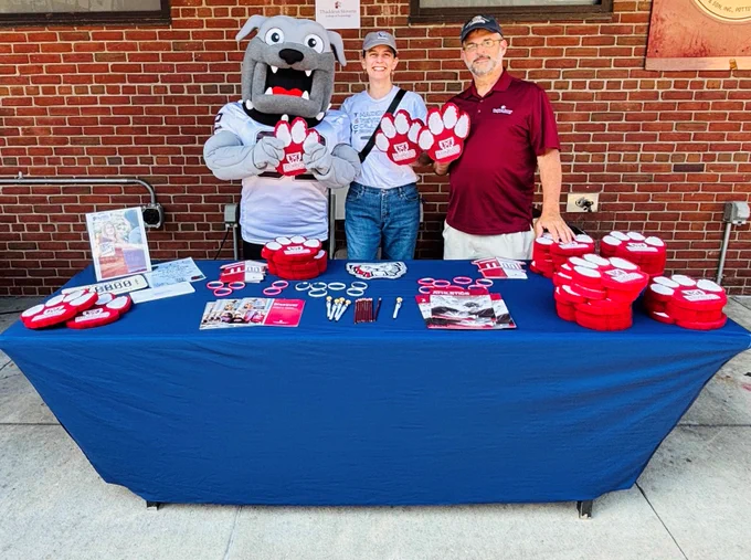 Three people, including a mascot in a bulldog costume, stand behind a table with red paw-shaped items and promotional materials against a brick wall.