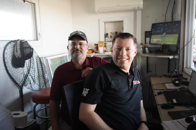 Two men sitting in a small, cluttered room and a TV showing a baseball game in the background.