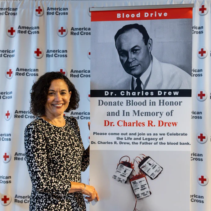 An individual stands next to a banner at an American Red Cross event. The banner features an image of Dr. Charles R. Drew and text about donating blood in his honor.