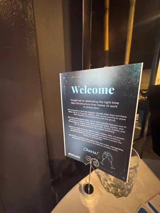 A welcome sign on a table introduces a celebration for right-time data infrastructure in AI production, mentioning Estuary's new Agent Skills and unlimited throughput updates. The sign includes an illustration of clinking champagne glasses.