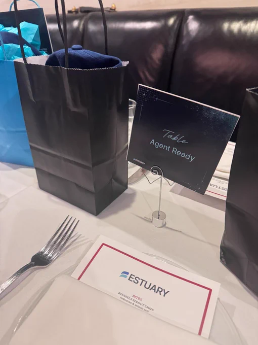 A dining table at a restaurant is set with a white tablecloth, black gift bags, and a sign holder displaying "Table Agent Ready." A name card on the table reads "ESTUARY." A fork is placed to the left of a white plate, and a blue bag is visible in the background.