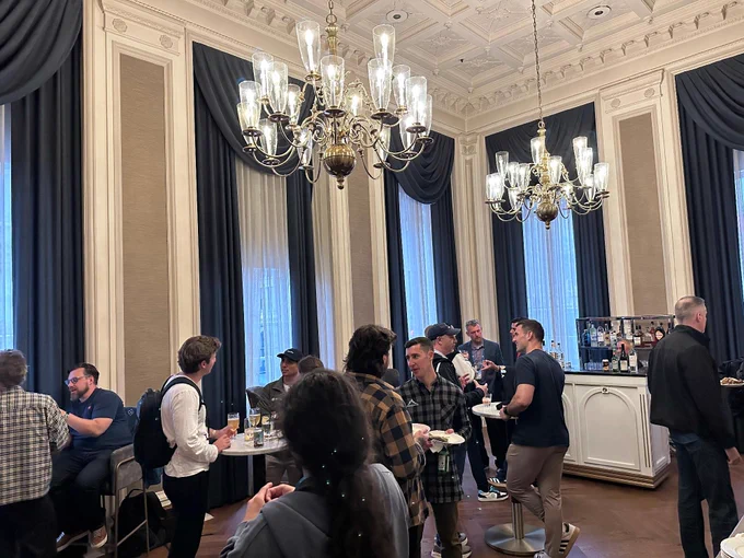A social gathering in a large, elegant room with chandeliers, tall windows, and curtains. People are mingling and chatting near a white bar with various beverages. The atmosphere appears lively and relaxed.