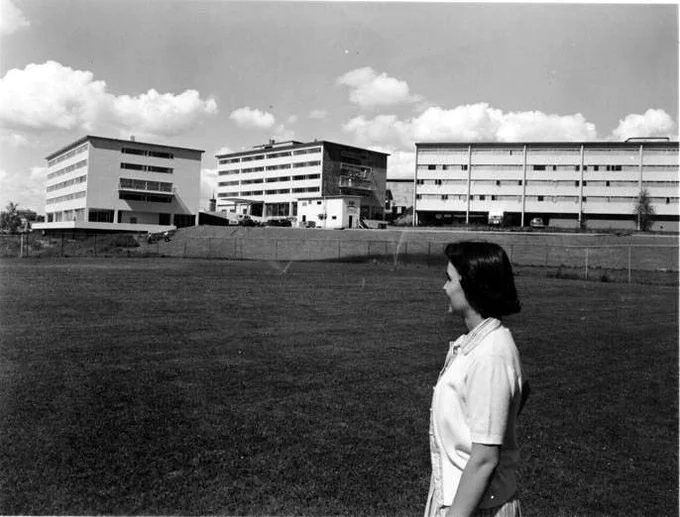 Black-and-white photograph showing a wide, open grassy field in the foreground, with a single person standing in profile on the right side of the image, facing toward a group of campus buildings in the distance. The person stands upright with arms at their sides and short, dark hair, wearing a light-colored collared shirt. Beyond the field, a row of mid-century institutional buildings stretches across the background: three rectangular, multi-story structures with flat roofs and long horizontal rows of evenly spaced windows. One building at center has a slightly different facade with a recessed or darker section. A low retaining wall or slope separates the field from the buildings, and a few small vehicles are faintly visible near the structures. The sky above is bright with scattered, puffy clouds, providing strong contrast to the dark tones of the buildings and field.
