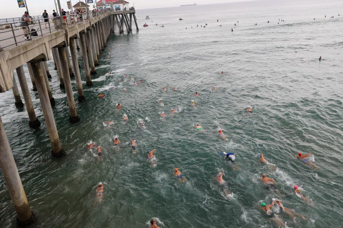 Swimmers participate in an ocean event near a pier at Huntington Beach. The water is filled with people wearing colorful swim caps, and the horizon shows a few ships. The pier is lined with spectators and flags.
