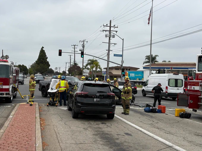 Firefighters, paramedics, and law enforcement personnel work at the scene of a multi vehicle traffic collision at a signalized intersection. A black Nissan Rogue is stopped in the foreground while emergency crews assess damaged vehicles and prepare patient transport. Fire engines, rescue equipment, and medical gear are positioned throughout the roadway under overcast skies as responders coordinate incident operations