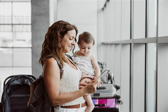 A person holding a child stands next to luggage by large airport windows. The person is looking at a smartphone screen.