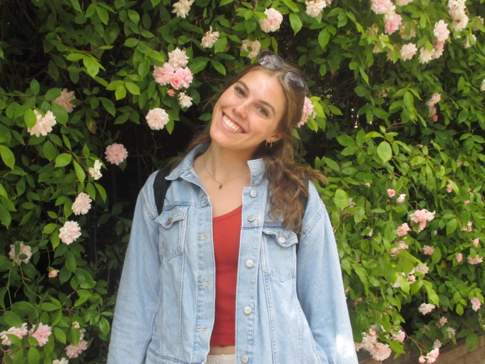A person is smiling in front of a lush wall of greenery adorned with pink flowers. The person is wearing a denim jacket and sunglasses are atop their head.