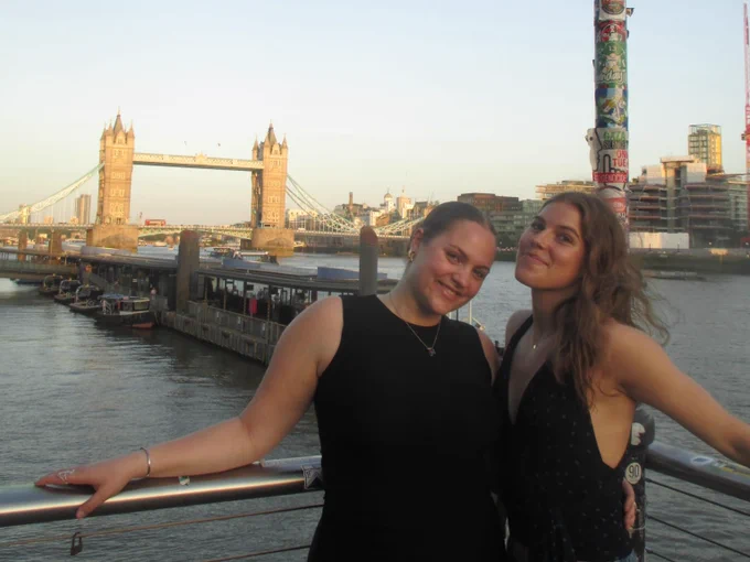 Two individuals are smiling and standing in front of the River Thames with Tower Bridge in the background. There is a pole covered with stickers next to them. The scene is during daylight with a clear sky.