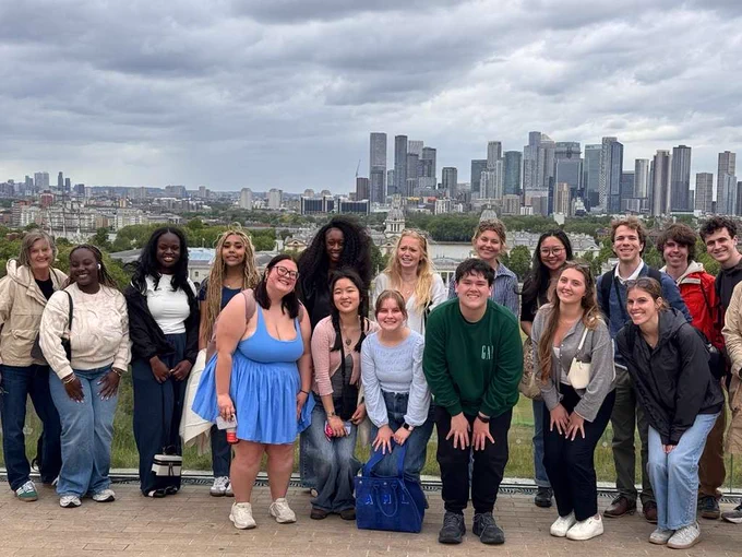A group of people are posing for a photo on an elevated walkway with a cityscape in the background. Cloudy skies are overhead, and the skyline includes iconic buildings such as The O2 in London. The group appears happy and relaxed.