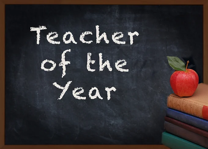 Written in white chalk on a blackboard are the words "Teacher of the Year." There is an apple on a stack of books in the foreground.