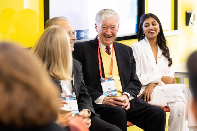Peter Cameron, a man in a suit & yellow sweater, wearing a \"GLOBAL LEADERSHIP Speaker\" badge, laughs. A young woman in a white embellished suit smiles beside him. Another person is partly visible, smiling. Background: \"26 SUMMIT\".