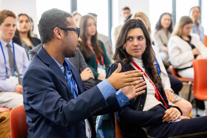 A man in a blue suit jacket and glasses gestures with open hands while speaking to a woman with long dark hair, wearing a dark jacket and white turtleneck. Both are seated in a lively discussion, with other blurred individuals in the background. The man's lanyard says \"Northeastern University\".