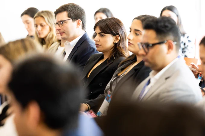 A group of diverse attendees seated in rows at a conference or event. A woman with dark hair wearing a black blazer and a clover earring is in profile, looking forward attentively. Next to her, a man in glasses and a dark suit jacket also looks forward. Other blurred faces fill the background.