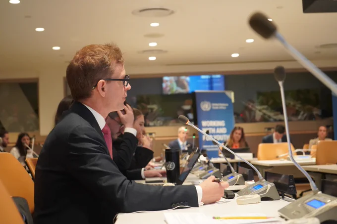 A participant in glasses speaks into a microphone during the meeting, with other delegates and the UN Youth Office banner visible in the background.
