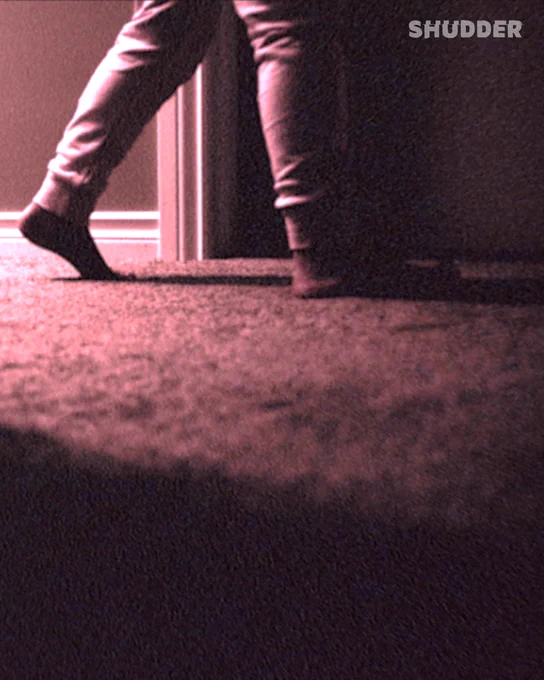 A moody, low-light scene of a person walking in a dimly lit hallway. The image has a grainy texture, capturing a sense of mystery and suspense.