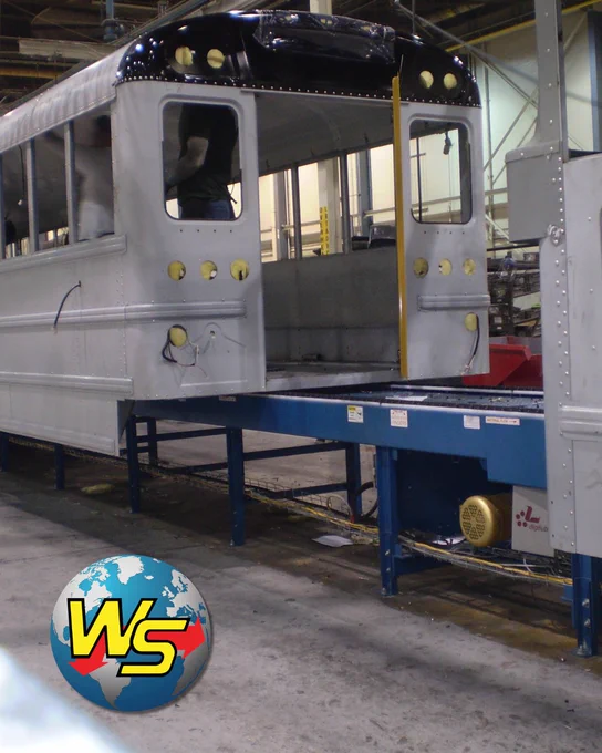 A large bus body in mid-assembly sits on a Webb-Stiles dual strand chain conveyor inside a manufacturing facility. The blue conveyor structure is visible beneath and alongside the bus, with the Webb-Stiles logo in the lower left corner.