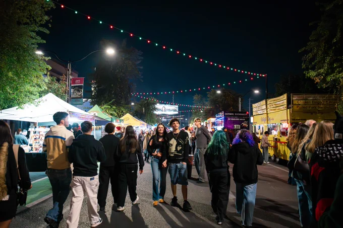 A lively street market scene at night with people walking under string lights. Food stalls and vendor tents line the street, including brightly lit yellow food trucks.