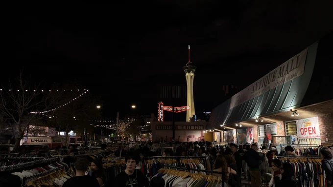 A bustling nighttime street market with numerous people browsing clothing racks. In the background, the illuminated Stratosphere Tower is visible, and nearby buildings display neon signs. String lights are strung overhead.