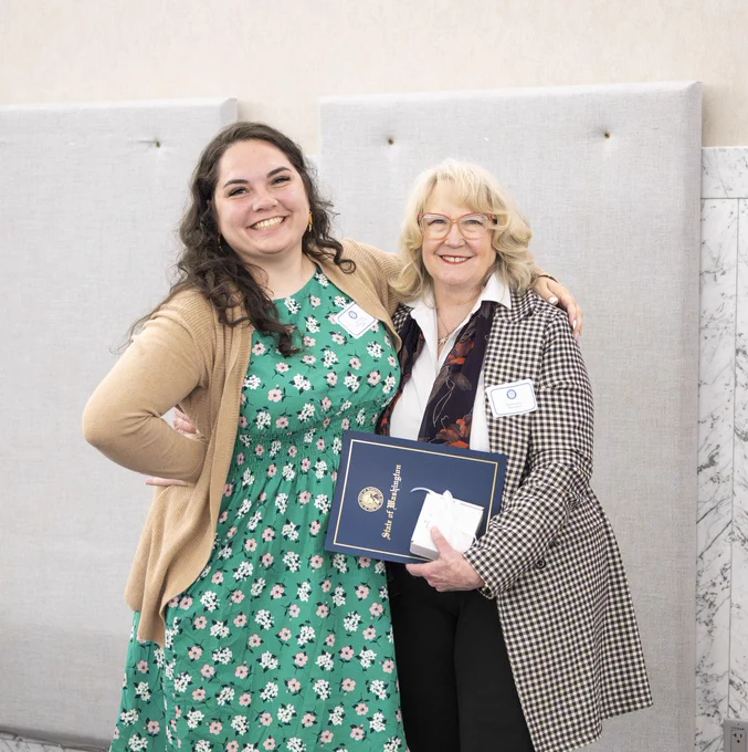 Two women standing together, one holding a certificate folder, both wearing name tags and smiling in a formal setting.