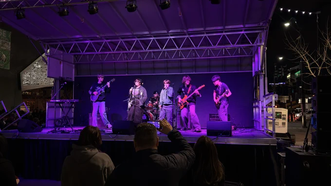 A band is performing on an outdoor stage under purple lighting. Several musicians are playing instruments, including guitars and a drum set. An audience is gathered in front, with one person holding up a smartphone to record the performance. It is nighttime, and the stage is set in a city environment with visible buildings and streetlights.