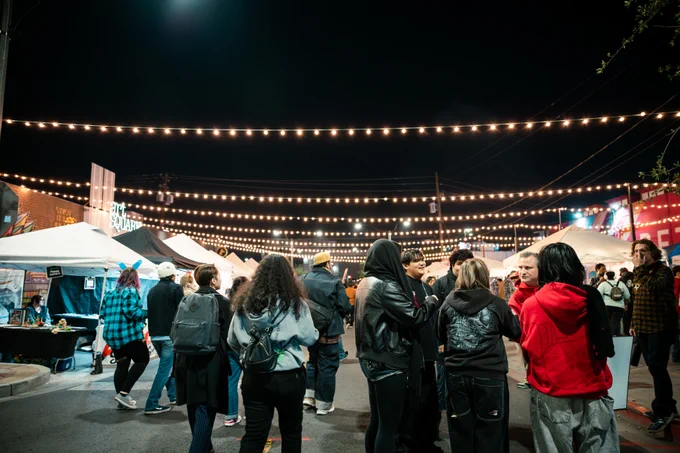 Outdoor night market with people gathered under strings of lights. Stalls line the street, and there are various booths. The scene is lively with groups socializing.