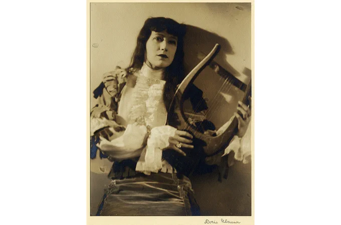 A person dressed in elaborate, ruffled attire poses holding a lyre.