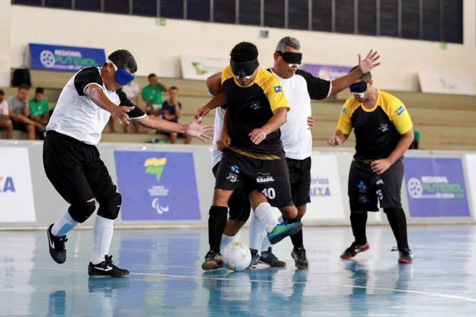Atletas da UNIACE-DF e da ADVP-PA disputam a bola durante partida válida pelo Regional Centro-Norte de Futebol de Cegos. No centro da imagem, um jogador da equipe do Distrito Federal, com uniforme preto e amarelo, tenta proteger a posse de bola enquanto é cercado por adversários de uniforme branco e preto. Todos os atletas utilizam vendas nos olhos e mantêm os braços abertos para orientação durante a jogada. Ao fundo, aparecem as arquibancadas e as placas de divulgação da competição.