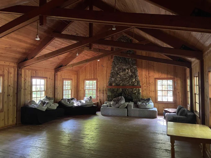 open room with wood paneling and friezes, a gray stone fireplace in the back. there are black and gray couches with gray pillows around. the flooring is wood and there are windows on the walls