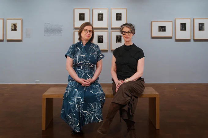 Two individuals are sitting on a bench in an art gallery, with framed photographs displayed on the wall behind them. The gallery has a calm atmosphere, with wooden flooring and soft lighting.