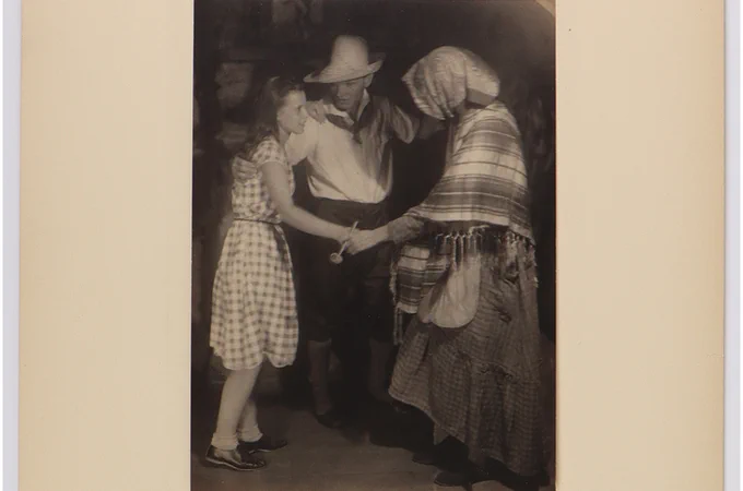 A person in a checkered dress and another in a wide-brimmed hat dance with an individual in a shawl and skirt. 