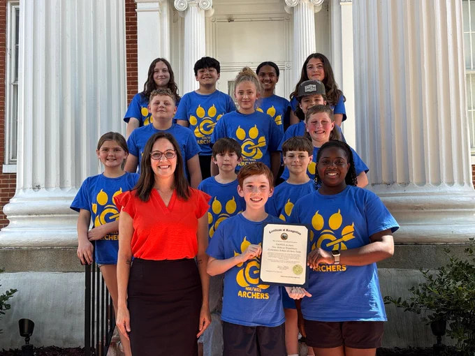 A group of children and one adult stand together on the front steps of a historic brick building with large white columns. The children are wearing matching bright blue T‑shirts featuring a yellow paw-print logo and the word “ARCHERS” printed beneath it. They are arranged in multiple rows on the steps, creating a tiered formation. One child at the front holds a framed certificate of recognition with an official seal at the bottom. The adult, positioned at the front left of the group, is wearing a bright orange top and a dark skirt. The background features tall, fluted white columns, decorative molding, and part of a large wooden doorway. 