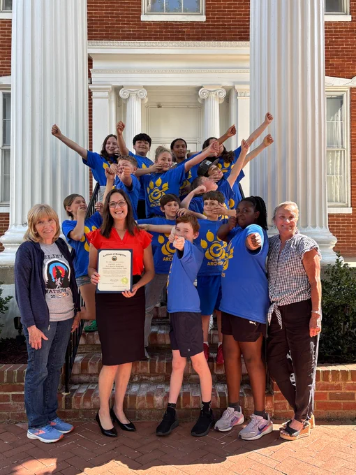 A large group of children and several adults gather on the brick steps of a historic building with tall white columns and red brick walls. Most of the children wear matching bright blue shirts featuring a yellow paw-print logo and the word “ARCHERS.” The group is arranged in an energetic, celebratory pose, with many children raising their arms upward or outward. At the center front, an adult wearing a bright orange top and a dark skirt holds a framed certificate. One child stands directly in front of the certificate holder, partially leaning toward the group. Two additional adults stand at the outer edges of the group, one on each side.