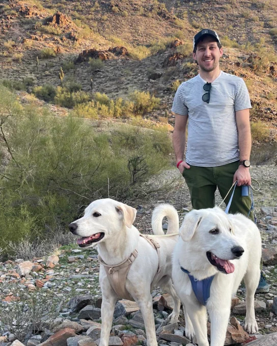 A person (Noah) stands with two white dogs on a rocky, desert trail with sparse vegetation and cacti on a hillside in the background.