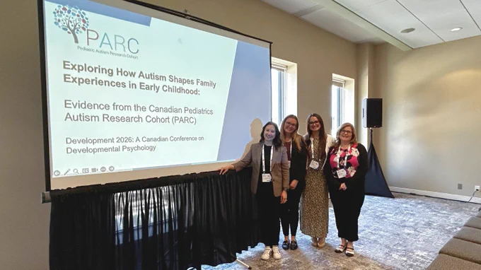 (Left to right) Dr. Vivian Lee, Melissa Susko, Renee Baysarowich, and Jennifer Crookshank stand and smile next to a large projector screen with a PowerPoint title slide with text, "Exploring How Autism Shapes Family Experiences in Early Childhood: Evidence from the Canadian Pediatrics Autism Research Cohort (PARC), Development 2026: A Canadian Conference on Developmental Psychology".