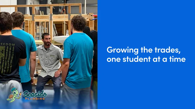 A group of people gather in a workshop setting, listening to an individual speaking. The environment features various plumbing equipment. The left corner displays the Seaside Plumbing logo, and a large blue panel on the right contains the text: "Growing the trades, one student at a time."