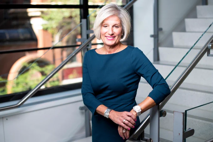 A smiling older woman with short blonde hair and bright pink lipstick, wearing a dark teal dress. She leans on a modern metal and glass staircase railing, with a blurred window showing greenery and a building in the background.