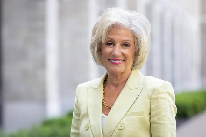 A smiling woman with voluminous blonde/white hair looks directly ahead. She wears a light green blazer over a white top with a chunky gold chain necklace. A blurry light-colored building and green foliage are in the background.