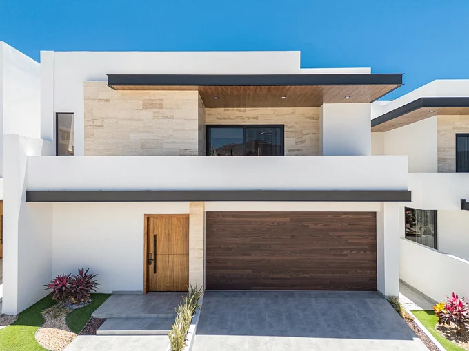 A modern, two-story house with a flat roof and a large garage. The exterior features white walls with stone accents and a wood tone garage door. There are small landscaped areas with plants on either side of the entrance.
