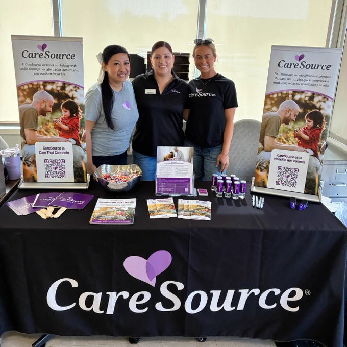 Three people stand behind a table with a CareSource tablecloth, featuring promotional materials. Two CareSource banners with images and text stand behind them.