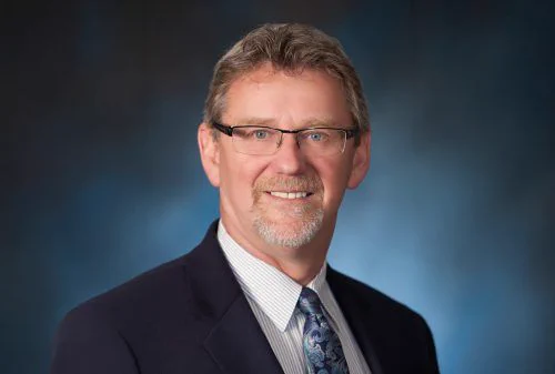Portrait of a smiling man in a suit with glasses, short hair, and a beard. Blue gradient background. Professional and approachable tone.