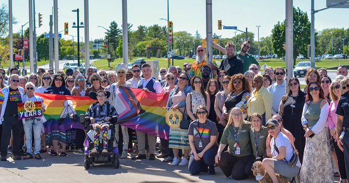 A large group of people is gathered outdoors holding the Pride and Two-Spirit flags.