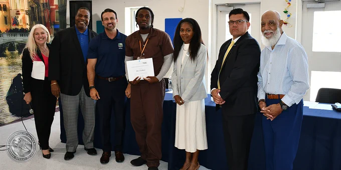 A group of seven people stand indoors, with one person holding a certificate. They are dressed in business and casual attire, and appear to be at an event or ceremony. The setting includes a table with a blue cloth. 