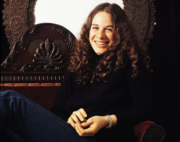 Carole King, joven, vestida con vaqueros y jersey negro, sentada sonriente en un sillón de madera muy señorial. Discogs.