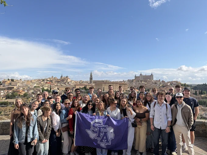 A group of student gathered outdoors in front of a scenic cityscape. They are holding a purple flag with the letters "SFA" on it. The sky is clear with some clouds. The background features historic buildings and a wide view of the city.