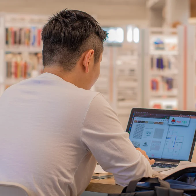 A person in a white shirt is sitting at a desk in a library, working on a laptop. Books are visible on the shelves in the background. 