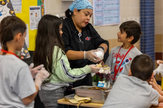 A Castleberry ISD chef is wearing a blue bandana and demonstrating a cooking technique to a group of students gathered around a table. The children are wearing gloves and observing closely.
