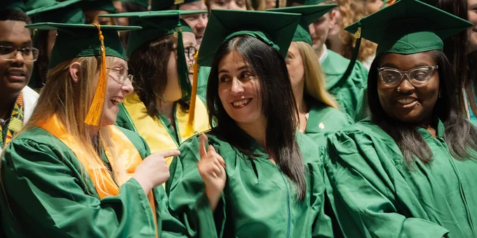 Group of FCC graduates in green caps and gowns sitting together during a ceremony, engaging in conversation and celebration.