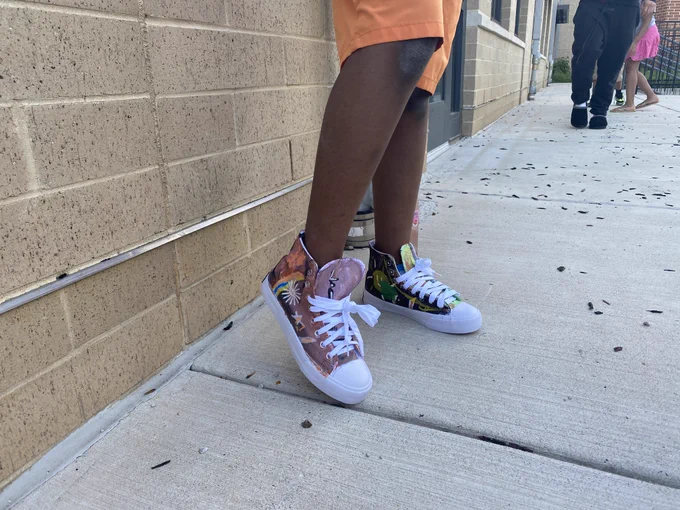 Person wearing colorful high-top sneakers with white laces standing on a concrete sidewalk next to a brick wall.